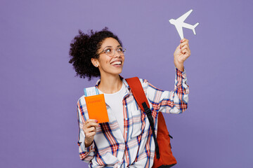 Young tourist girl woman of African American ethnicity student 20s in shirt backpack travel abroad hold passport ticket airplane mockup isolated on plain purple background. Air flight journey concept.
