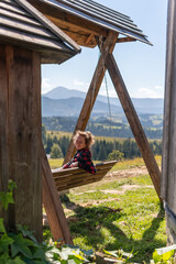 A young woman is sitting and enjoying time, relaxing on a wooden swing in the mountains on a bright summer sunny day