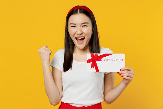 Vivid young girl woman of Asian ethnicity 20s years old wear white t-shirt hold gift certificate coupon voucher card for store doing winner gesture isolated on plain yellow background studio portrait
