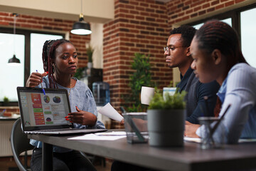 Marketing company team leader presenting on laptop business analytics chart. African american businesswoman introducing to colleagues new management and business plan while in modern office.