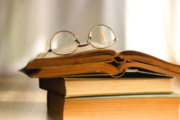 Open book and reading glasses on the table, illuminated by sunlight. Stack of vintage books in the background. Selective focus.