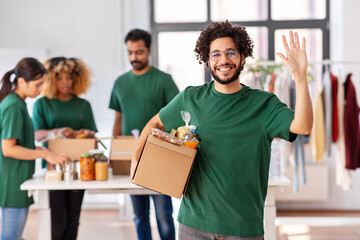 charity, donation and volunteering concept - happy smiling male volunteer with food in box waving hand over and international group of people at distribution or refugee assistance center