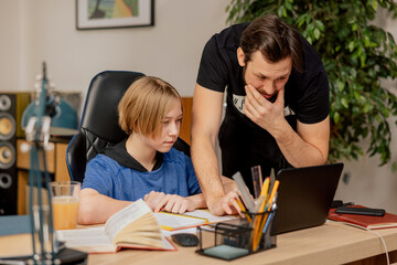 Fototapeta na wymiar A caring father tries to help son studying at home remote learn solve technical problems on laptop, the boy sits at desk prepared for e-learning with a notebook and a book.