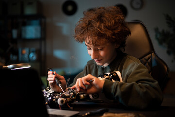 A child with curly hair sits at a desk working on soldering a robot, fixing wires, cables, toy electronics.