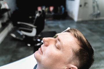 Visit to barbershop.Wax depilation,hair removal between the eyebrows.Stylish man makes fashionable haircut.Barber,hairdresser,stylist.Client,customer sits in dark men's beauty salon covered with cape