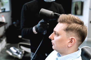 Visit to barbershop. Stylish man makes fashionable haircut. Barber,hairdresser,stylist with scissors, comb cutting,dryer.Combing hair.Client,customer sits in dark men's beauty salon covered with cape