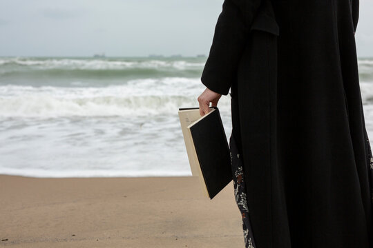 Plano de un libro no identificable con tapas negras sostenido en las manos de una mujer en la playa durante un d&iacute;a nublado. 