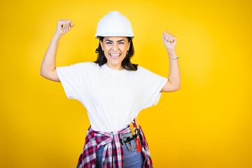 Young caucasian woman wearing hardhat and builder clothes over isolated yellow background very happy and excited making winner gesture with raised arms, smiling and screaming for success.