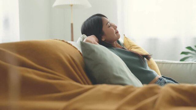 Relaxed young asian woman enjoying rest on comfortable sofa at home, calm attractive girl relaxing and breathing fresh air in home.