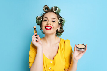 Comic portrait of young emotional girl, housewife with curlers on her head isolated on blue studio...