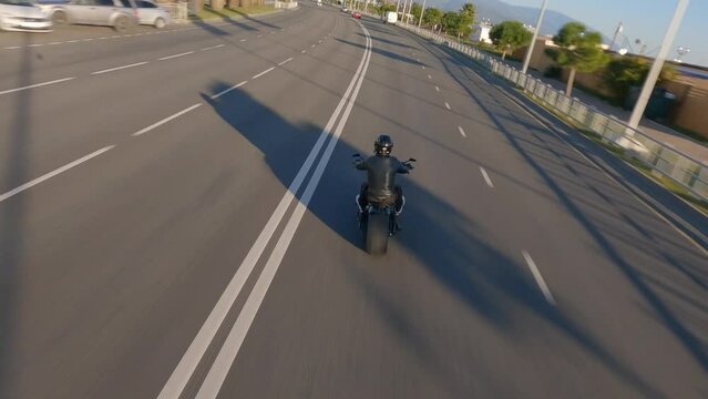 Black lether biker riding on custom bike at highway traffic urban neighborhood nature landscape. Flying over sunlight touristic city sea beach street district town panorama sports FPV drone shot