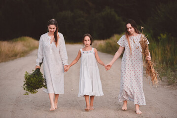 Three girls in long white nightgowns walk barefoot on a sandy road near a forest on a warm summer...