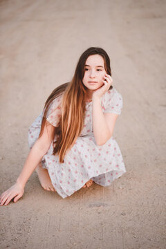A beautiful young girl with long hair in a white nightgown sits with her nose on a sandy road, looks away. Warm summer day, walk in the countryside.