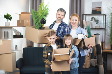 Happy Caucasian man and woman with their two kids standing at new apartment with lots of boxes around. Concept of mortgage, family and real estate.