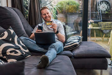 Smiling caucasian man, using his credit card and laptop, at home. - obrazy, fototapety, plakaty