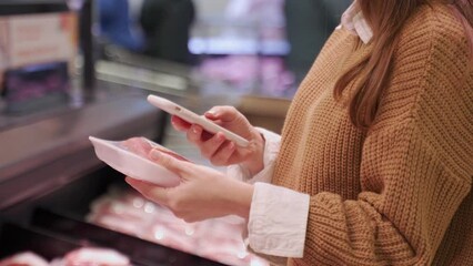 Young woman with a smartphone in her hand scans a package of meat in a supermarket