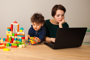 A happy mother works at a laptop and talks on the phone hugging her child who is playing next to a wooden construction set.