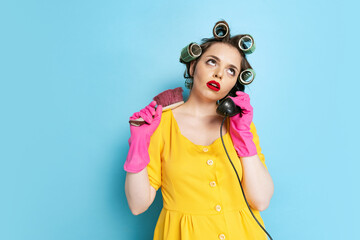 Young emotional girl, housewife with curlers on her head talking on retro phone isolated on blue...