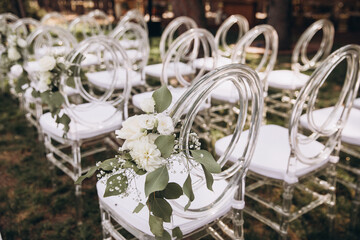 Wedding. Ceremony. There are white transparent designer chairs for guests on a green meadow in the forest. Chairs decorated with white flowers