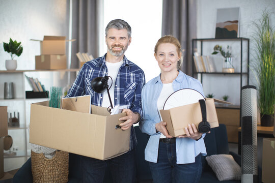 Caucasian middle aged couple in casual wear smiling and hugging at new flat, holding boxes with different decor and things. Young family moving to modern apartment. Cardboxes around.