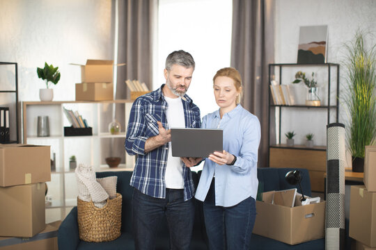 Caucasian mature couple standing among cardboxes and different things and using laptop, looking for the design and making online shopping. Family taking break during removing to new flat.