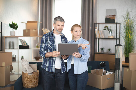 Caucasian mature couple standing among cardboxes and different things and using laptop, looking for the design and making online shopping. Family taking break during removing to new flat.