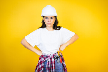 Young caucasian woman wearing hardhat and builder clothes over isolated yellow background skeptic and nervous, disapproving expression on face with arms in waist