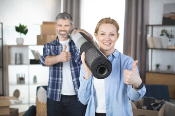 Smiling Caucasian couple carrying rolled rug after moving in a house. Focus on smiling pretty blond...