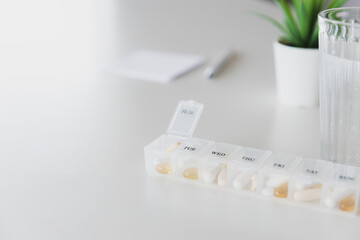 Closeup of medical pill box with doses of tablets for daily take a medicine with white, yellow drugs and capsules used for treatment, cure the disease. Glass cup of water on table, gray background.