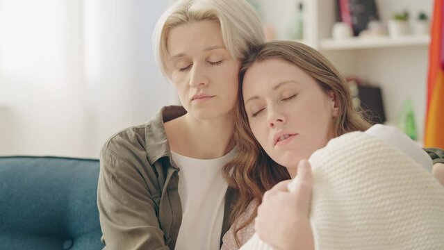 Exhausted Lesbian Couple Rocking Newborn Baby, Trying To Get Some Sleep On Sofa