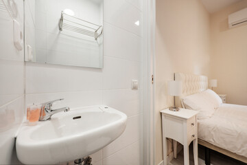 Bathroom with porcelain wall-hung sink, square frameless mirror, and bedroom with white bedding and white nightstands
