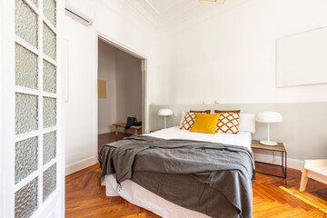 Bedroom with a large bed with many cushions, a gray blanket, twin bedside tables with white lamps and white woodwork