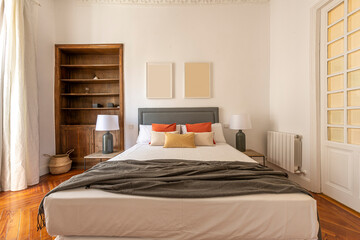 Bedroom with a large bed with lots of cushions, a gray blanket, two bedside tables with gray lamps with white shades and white joinery on doors and windows and a varnished pinewood floor