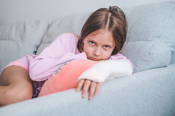 Close-up of a child's broken arm in a cast. The girl put her broken arm on the sofa and looks sadly at the camera. A child with a plaster on his arm.