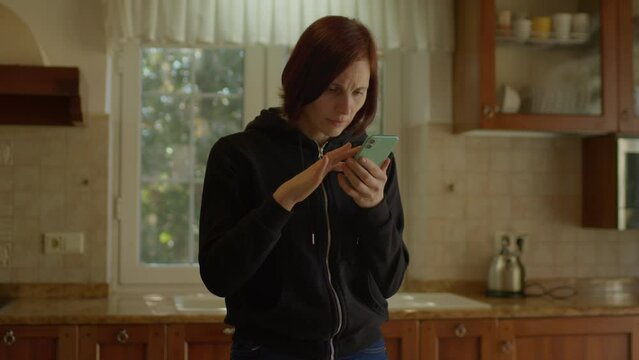 30s woman reading bad news on mobile phone and getting angry and nervous. Upset female using smartphone standing in the kitchen.