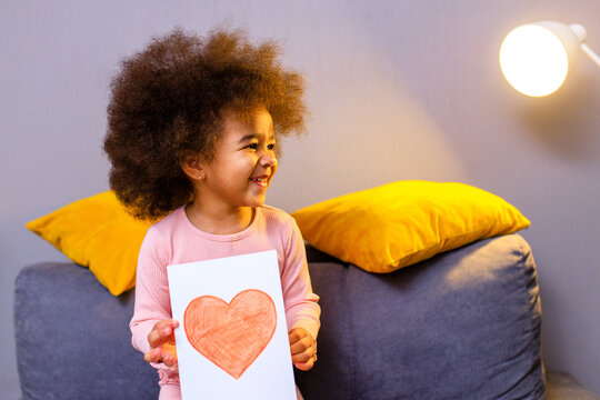 multicultural cute little curly girl holding post card with drawn heart in cozy living room at evening
