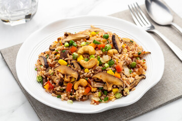 Vegetarian fried brown rice with cashews, shiitake mushrooms and colorful vegetables like onion, carrot, green pea, corn kernels and tomatoes. It is a healthy veggie dish in Thai food cooking style.