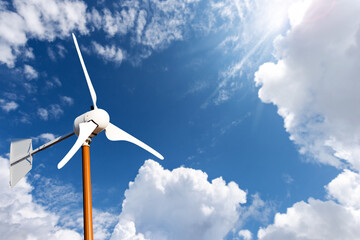Close-up of a small white wind turbine, against a clear blue sky with clouds, sunbeams and copy...