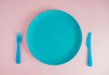 Blue empty plate with plastic knife and fork on a pastel pink color background. Flat lay minimal creative food concept with copy space. Breakfast, lunch, dinner. 