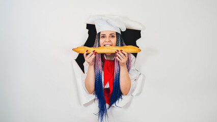 Young woman dressed as chef bites white bread, standing in hole of white background. Hungry female cook eating loaf in studio.