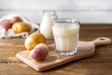 Potato milk in glass and milk jar on cutting board with raw potatoes in string bag. Wooden background. Rustic style