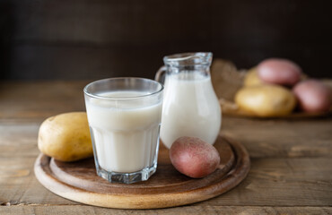 Vegan potato milk in glass and milk jar on cutting board with raw potatoes aside. Wooden background. Rustic style