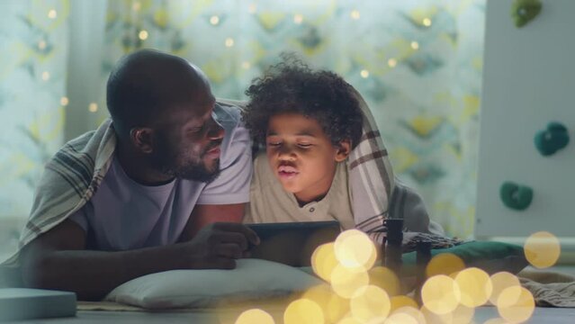 African American man discussing something on tablet with little son while lying together under blanket in cozy bedroom decorated with lights