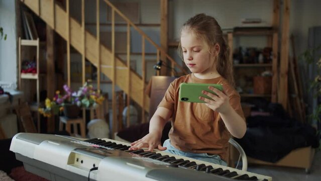 The girl watches a video lesson on the phone and tries to play the synthesizer