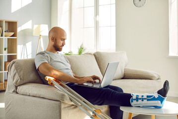 Unhealthy businessman with injured leg in bandage sit on sofa at home work online on computer....