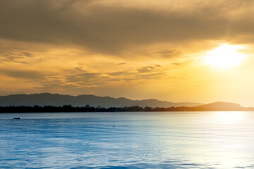 Abstract of wonderful light of the golden sky and the blue sea. with the silhouette of the forests and mountains on the distant shores. Sun shines out from the sky above.