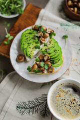 Open toasts with baked mini champignons, avocado slices, radish microgreens and cream cheese. Light healthy lunch in a plate on a board with a tablecloth. Minimalistic table setting.