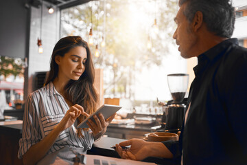 Processing payment. Cropped shot of an attractive young woman processing a male customers payment on her tablet while working in her coffee shop.