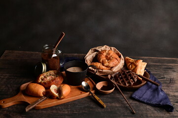 Breakfast Served with Croissants, ham and cheese bread, sausage buns, pies, waffles, pizza, coffee, and chocolate on wooden table with black background, selective focus.