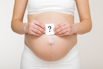 Hands holding card of question mark on young adult pregnant woman big naked belly. Guessing future...